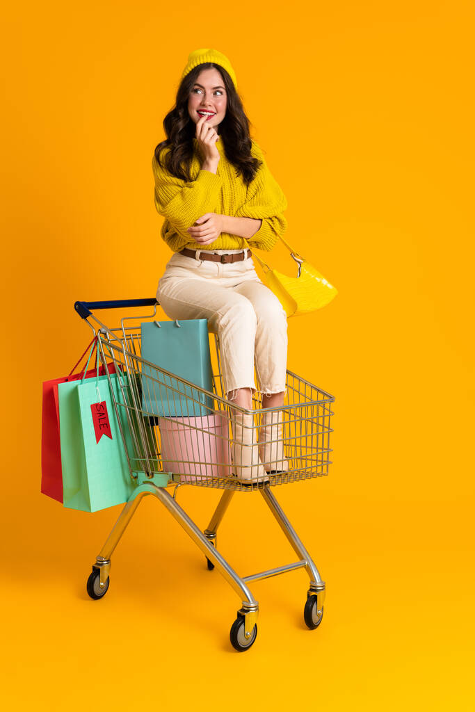 Weiße Frau lächelt, während sie im Einkaufswagen vor gelbem Hintergrund sitzt - Foto, Bild