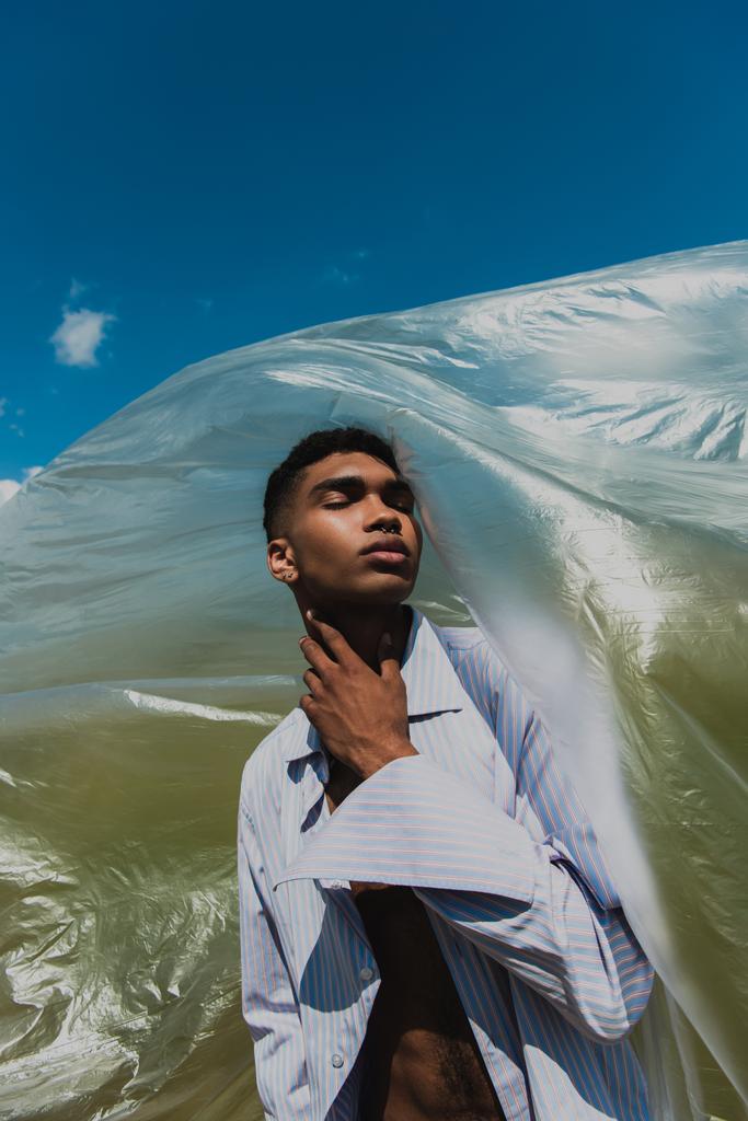 stylish african american man with closed eyes touching neck while posing near polyethylene under blue sky - Foto, imagen