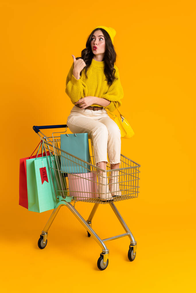 White woman pointing finger aside while sitting in shopping cart isolated over yellow background - Foto, immagini