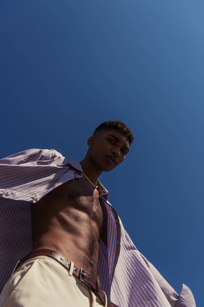 низький кут зору афроамериканця в смугастій сорочці, дивлячись на камеру на блакитне небо
 - Фото, зображення