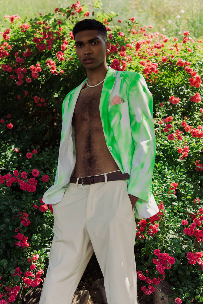 young and stylish african american man standing with hands in pockets near blossoming bushes - Foto, Bild
