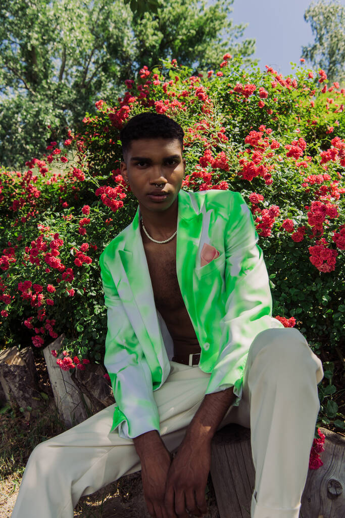 african american man in stylish blazer and beads sitting near blossoming bushes and looking at camera - Foto, afbeelding