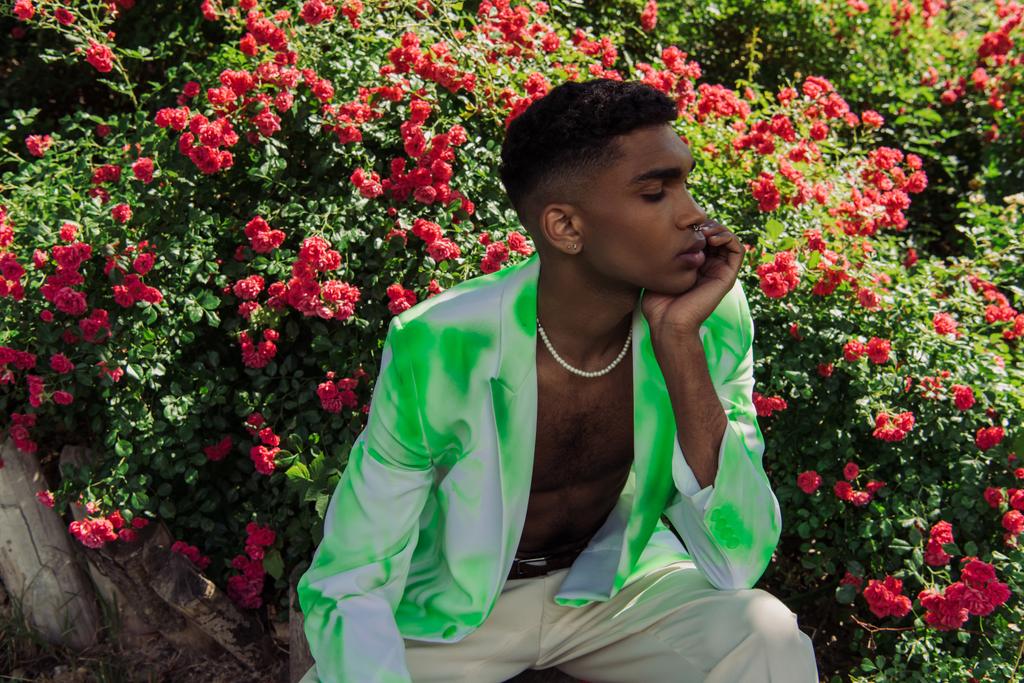 Parkta çiçek açan çalıların yanında oturan modaya uygun spor ceketli Afro-Amerikan genç adam. - Fotoğraf, Görsel