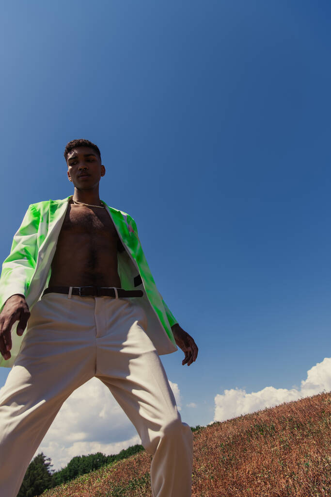 low angle view of african american man in white trousers and trendy blazer against blue sky - Фото, изображение