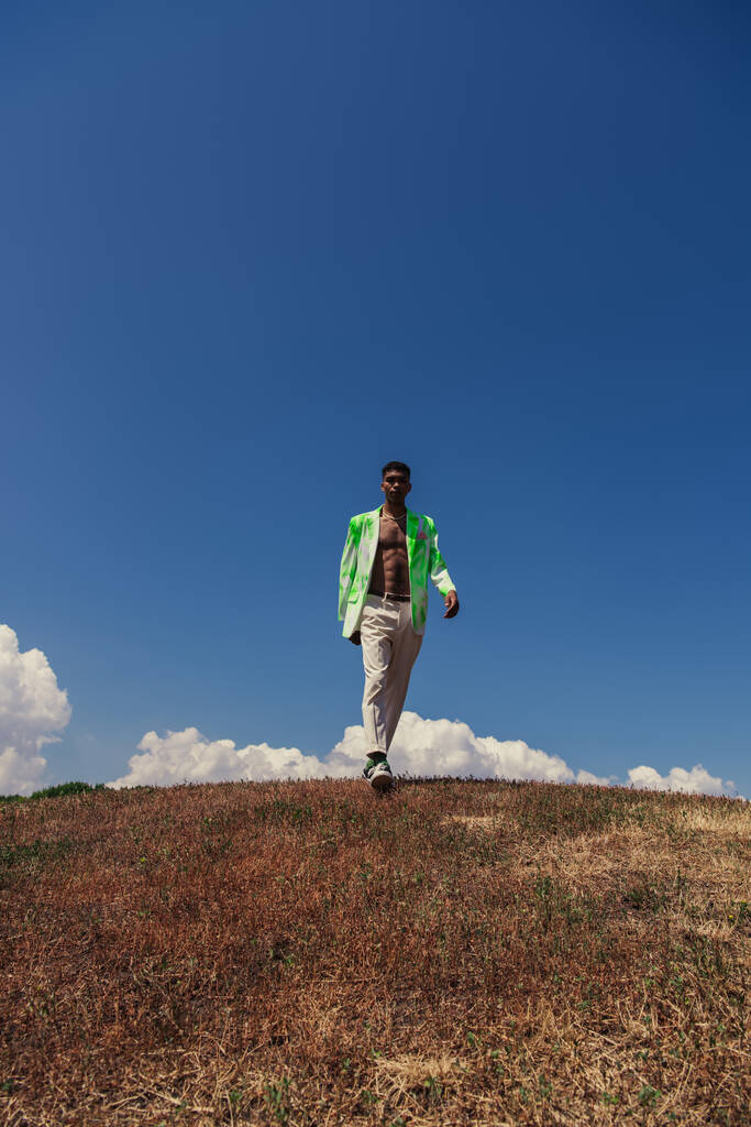 full length of african american man in trousers and blazer walking in field under blue sky with white clouds - Foto, immagini