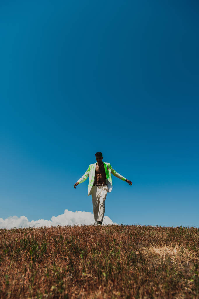 african american man in white trousers and blazer walking under blue sky in field - Photo, Image