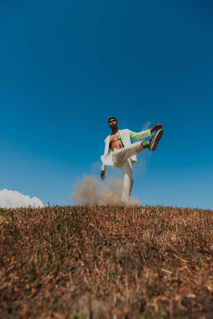 africký Američan ve stylovém oblečení pózuje se zvednutou nohou na louce pod modrou oblohou - Fotografie, Obrázek