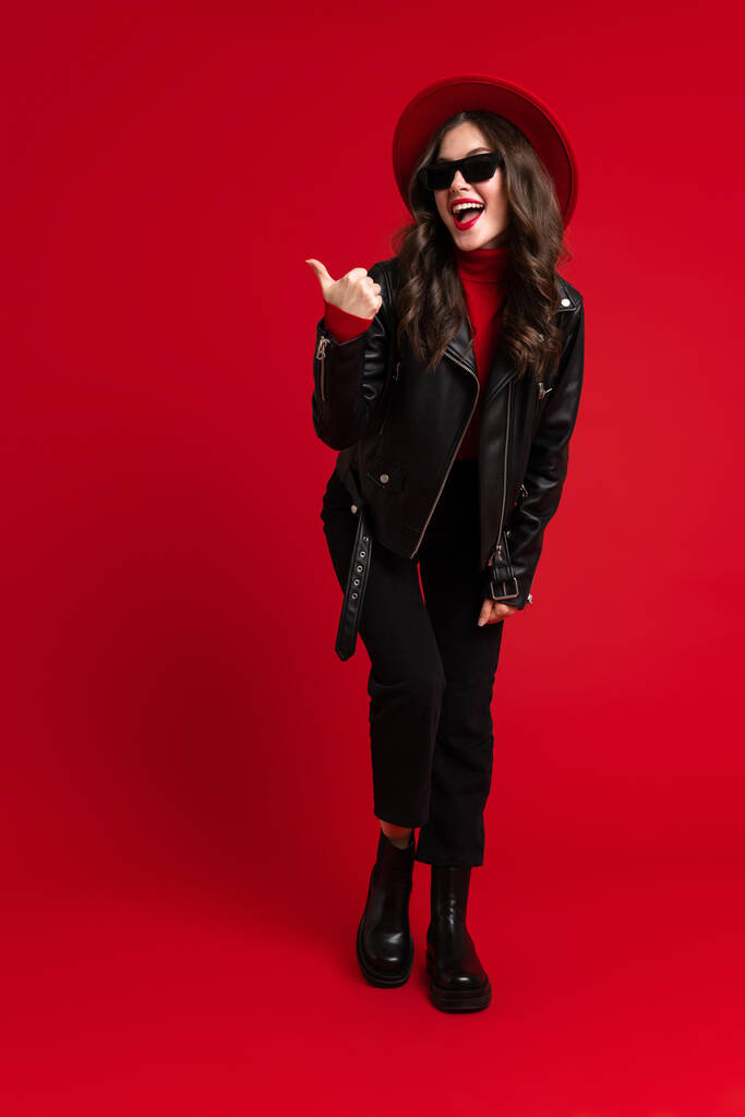 White woman in leather jacket smiling and pointing finger aside isolated over red background - Fotó, kép
