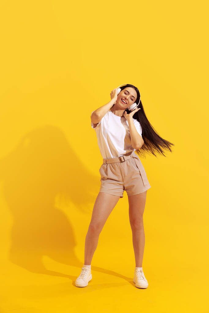 Soul music. Young charming girl with long dark hair in shorts and t-shirt isolated on bright yellow background. Concept of beauty, art, fashion, youth, health. Copyspace for ad - Fotó, kép