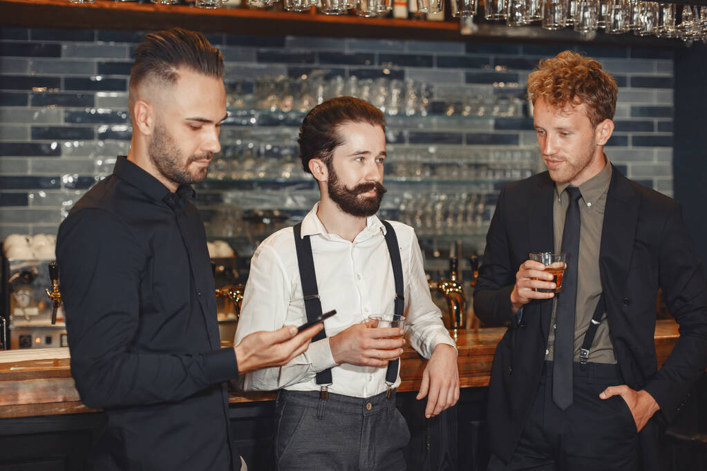 Meeting with the best friends. Three happy young men in casual wear talking and drinking beer while sitting in bar together. Man holding a phone in his hands. - Foto, Imagen