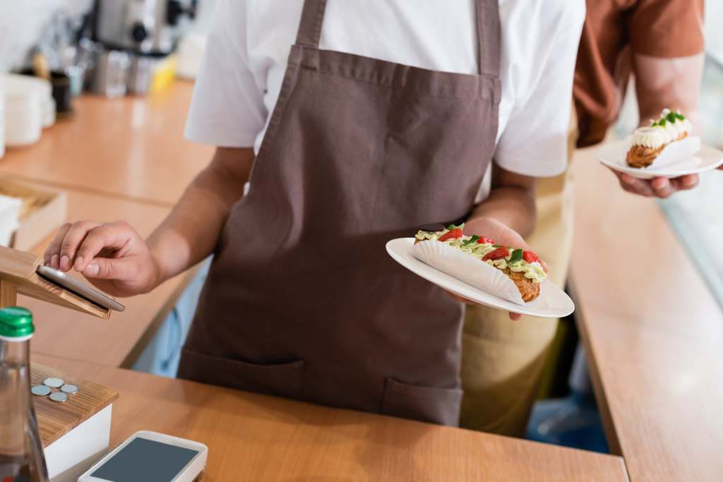 Обрізаний вид афроамериканського продавця, який тримає десерт і використовує цифровий планшет біля колеги в кондитерських виробах
 - Фото, зображення