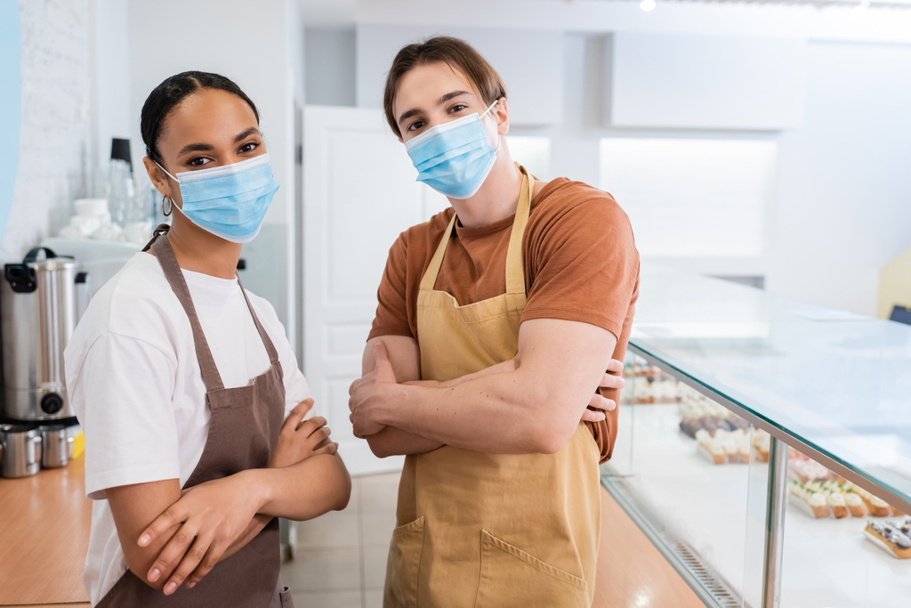 Interracial sellers in medical masks crossing arms in confectionery  - 写真・画像