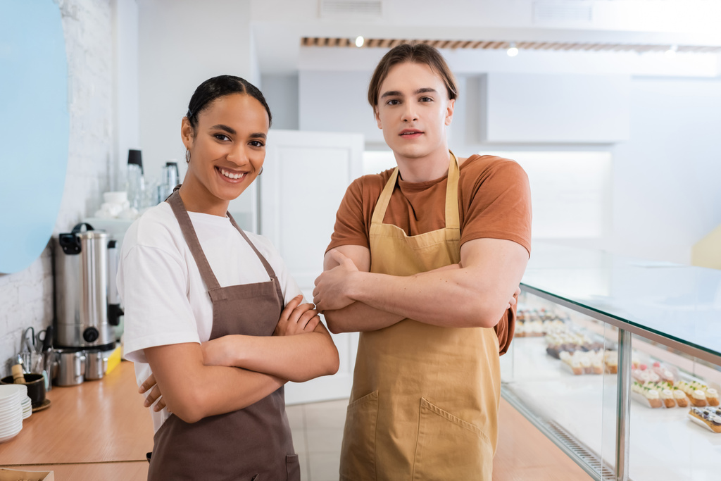 Smiling interracial sellers in aprons crossing arms in sweet shop  - Фото, изображение