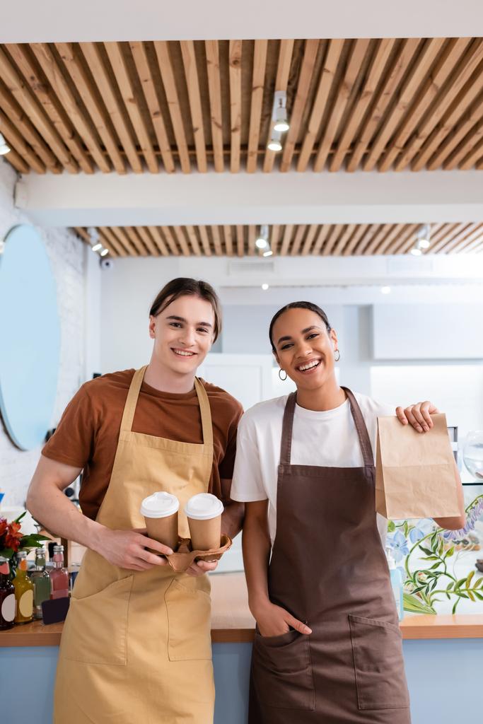Cheerful multiethnic sellers holding coffee to go and paper bag in confectionery  - Foto, Bild