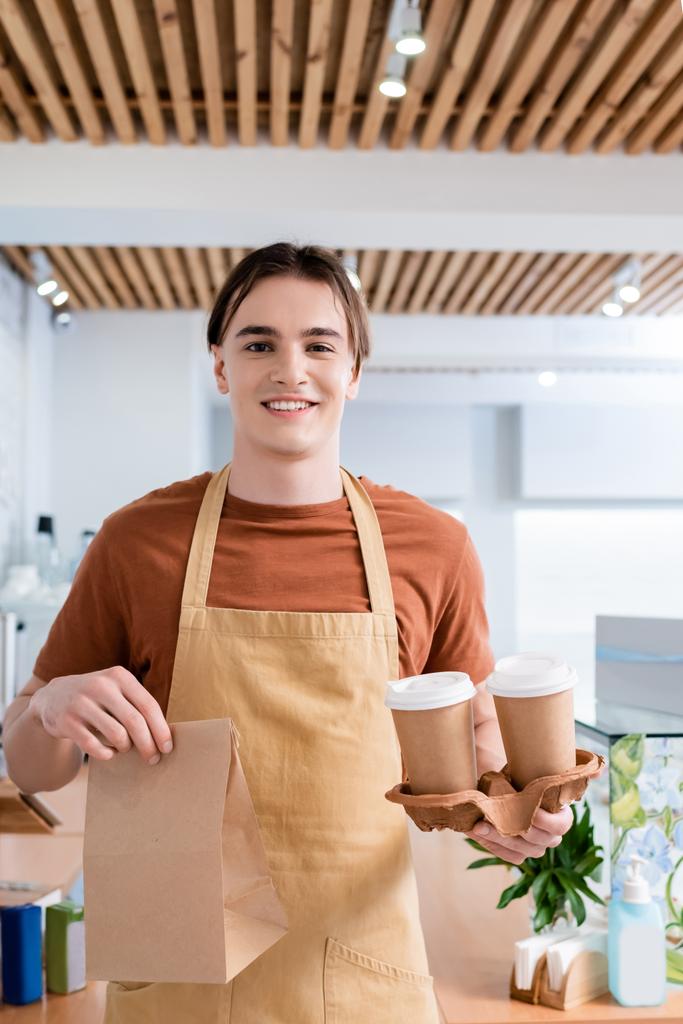Cheerful young salesman holding coffee to go and paper bag in sweet shop  - Photo, Image