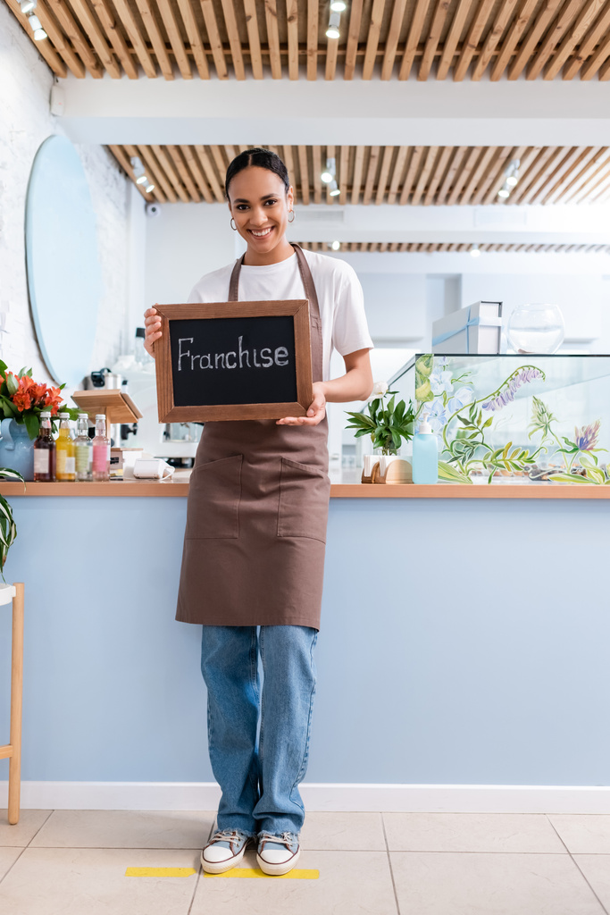 Positive african american seller holding chalkboard with franchise lettering in confectionery  - Foto, Bild