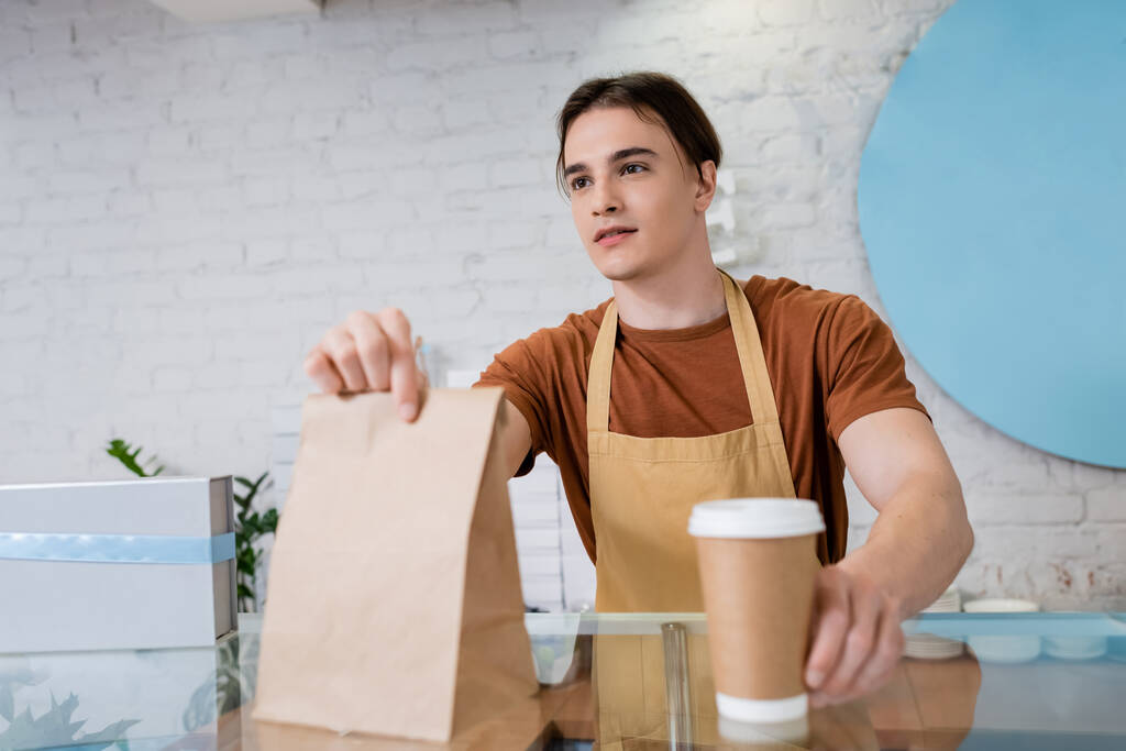 Seller in apron holding paper bag and takeaway drink in confectionery  - Φωτογραφία, εικόνα