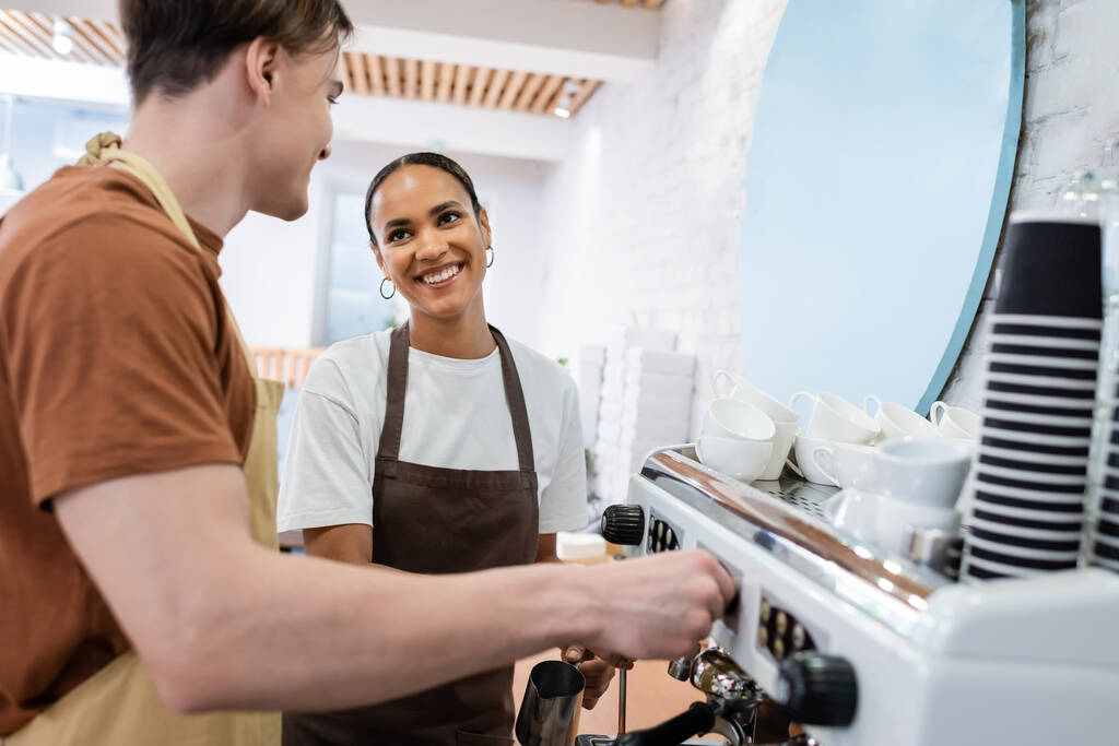 Lächelnder afrikanisch-amerikanischer Verkäufer mit Blick auf Barista, der Kaffee in Süßwaren kocht  - Foto, Bild