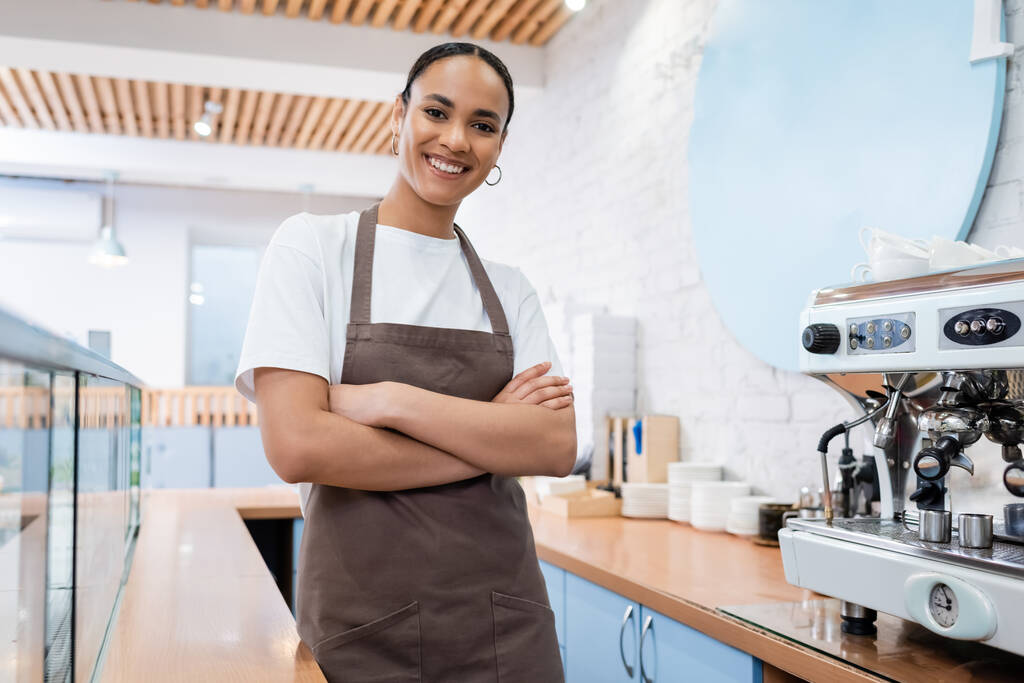 Smiling african american barista in apron crossing arms near coffee machine in sweet shop  - Foto, Imagen