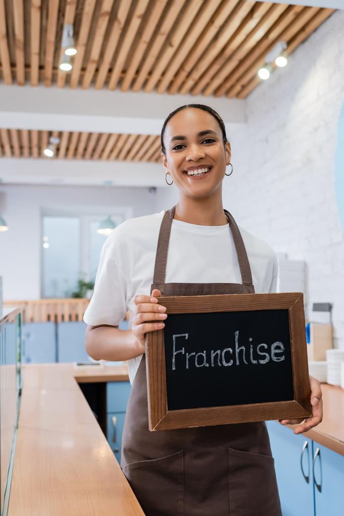 Positive african american barista holding chalkboard with franchise lettering in confectionery  - Foto, Imagem