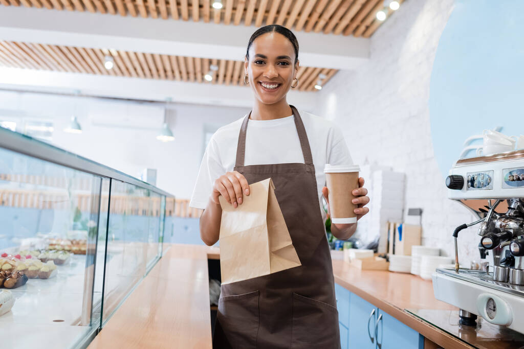 Positive african american saleswoman holding takeaway drink and paper bag in confectionery  - Φωτογραφία, εικόνα