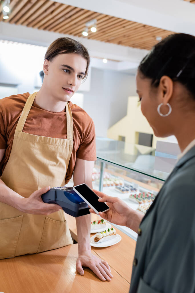 Young salesman holding payment terminal near african american customer with smartphone in confectionery  - 写真・画像