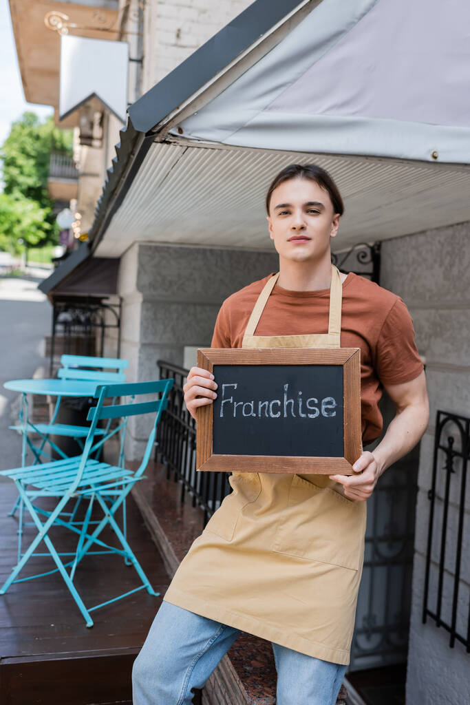 Young salesman holding chalkboard with franchise lettering on terrace of confectionery  - Foto, immagini