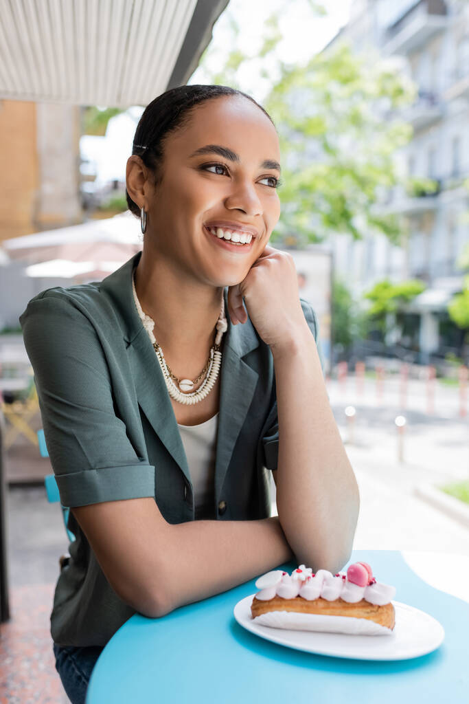 Усміхнений афроамериканський клієнт, що сидить біля смачного еклера на терасі кондитерських виробів
  - Фото, зображення