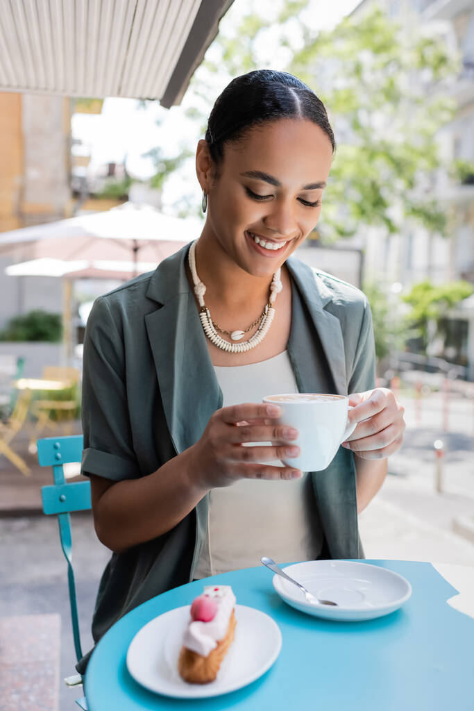 Positive african american woman holding cappuccino near blurred dessert on terrace of sweet shop  - Fotoğraf, Görsel