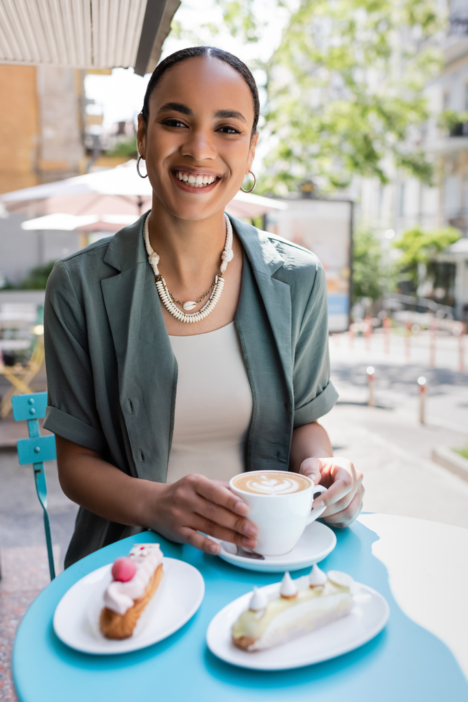 glimlachende Afro-Amerikaanse vrouw op zoek naar camera in de buurt van cappuccino en eclairs op het terras van zoetwaren  - Foto, afbeelding