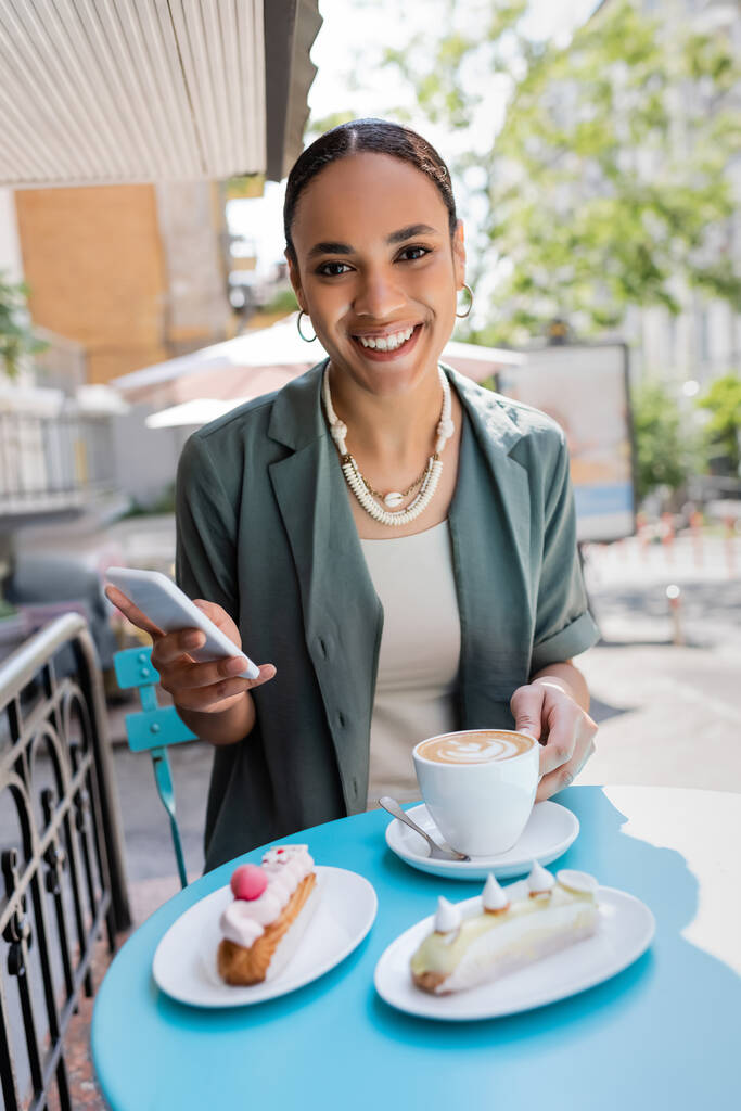 Pretty african american client holding smartphone near cappuccino and eclairs on terrace of confectionery  - Photo, image