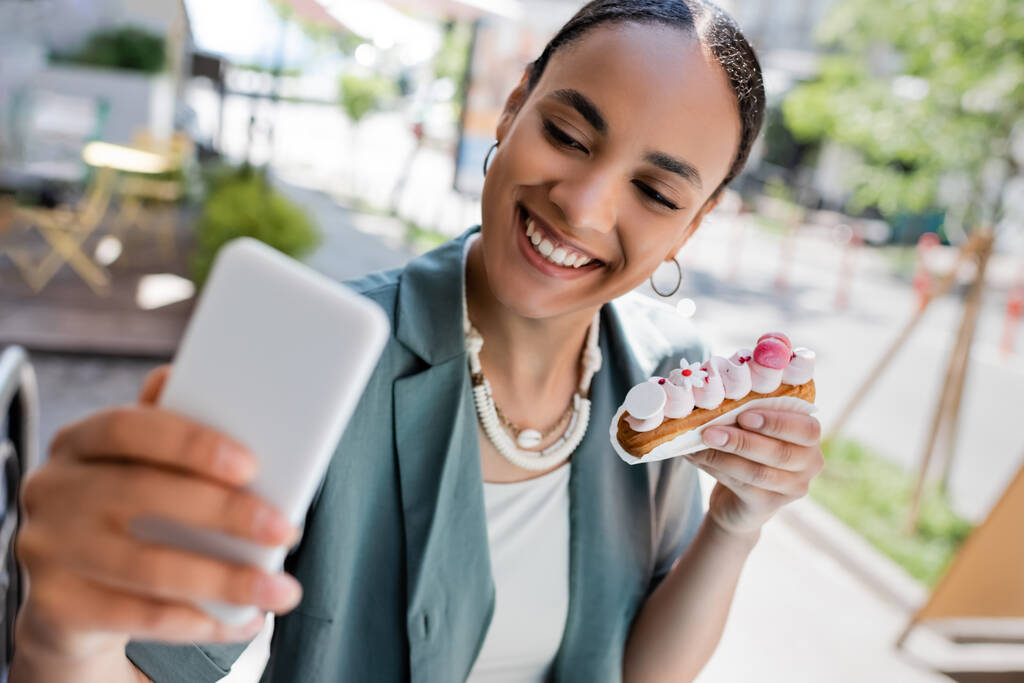 Iloinen afrikkalainen amerikkalainen nainen, jolla on eclair ja ottaa selfie älypuhelimella makeisten terassilla  - Valokuva, kuva