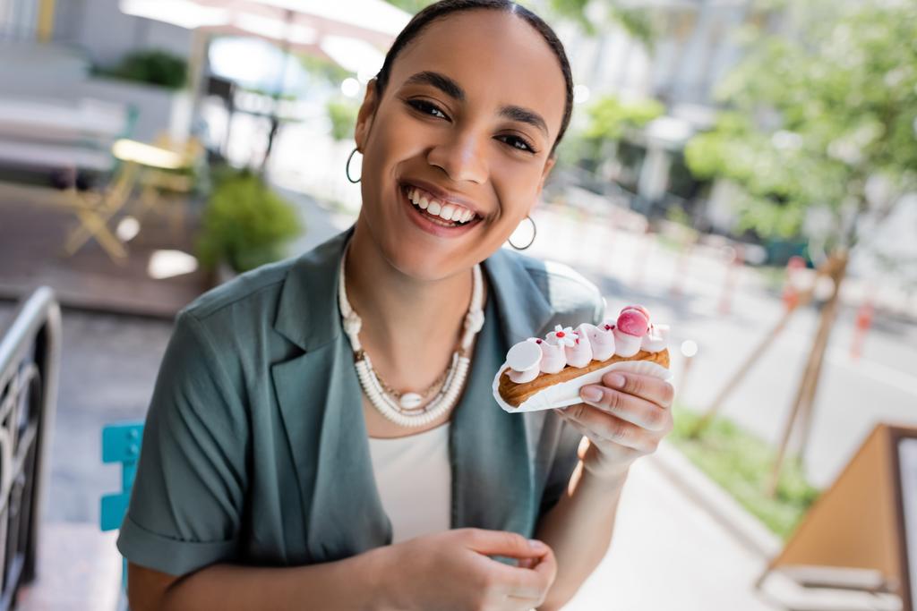 Cheerful african american woman holding eclair and looking at camera on terrace of cafe  - 写真・画像