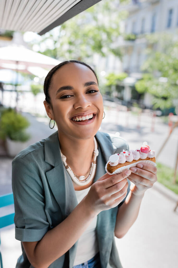 Positive african american client holding tasty eclair on terrace of cafe  - Foto, imagen