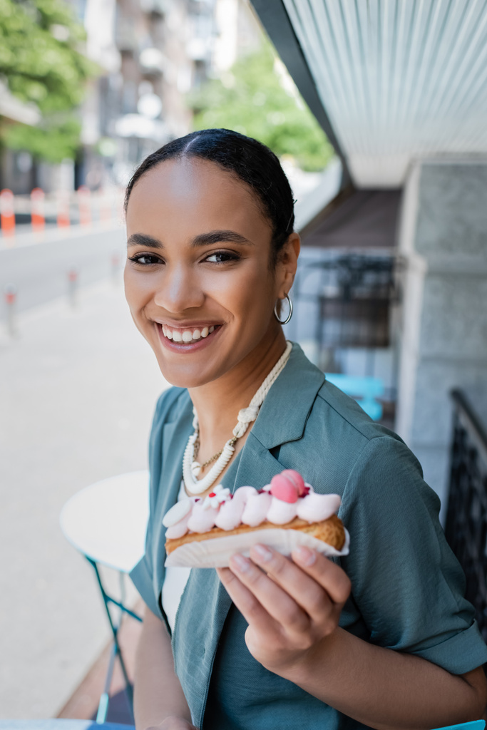 Positive african american customer holding blurred eclair on terrace of sweet shop  - 写真・画像