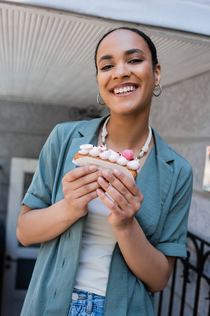 Smiling african american woman holding delicious eclair near sweet shop  - Photo, Image