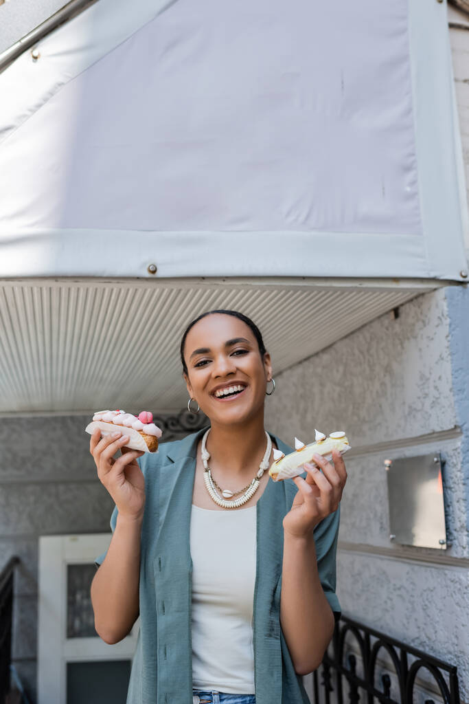 Happy african american woman holding tasty eclairs near sweet shop outdoors  - Foto, imagen