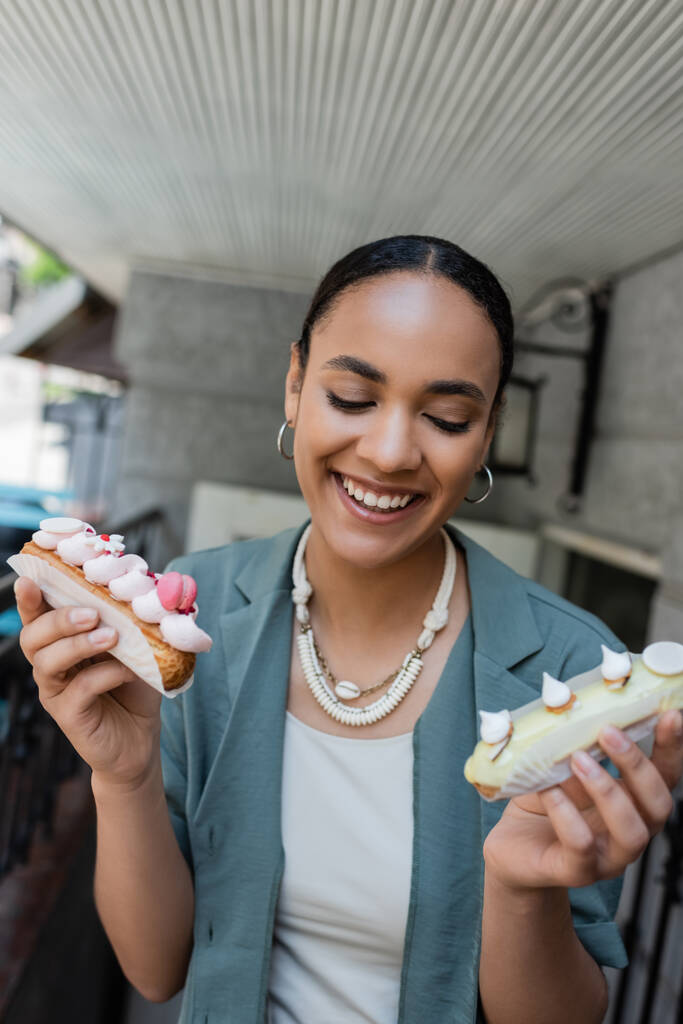Positive african american customer holding eclairs near confectionery outdoors  - Foto, imagen