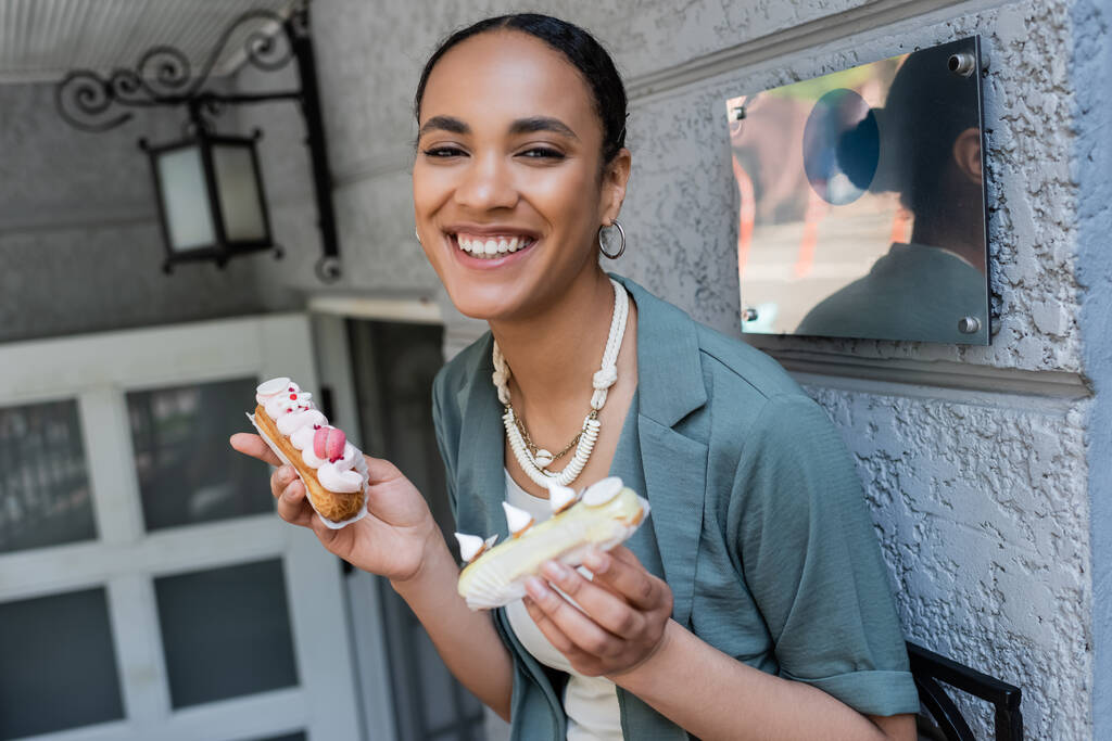 Pozitivní africký americký zákazník drží sladké eclairs v blízkosti cukrovinek venku  - Fotografie, Obrázek