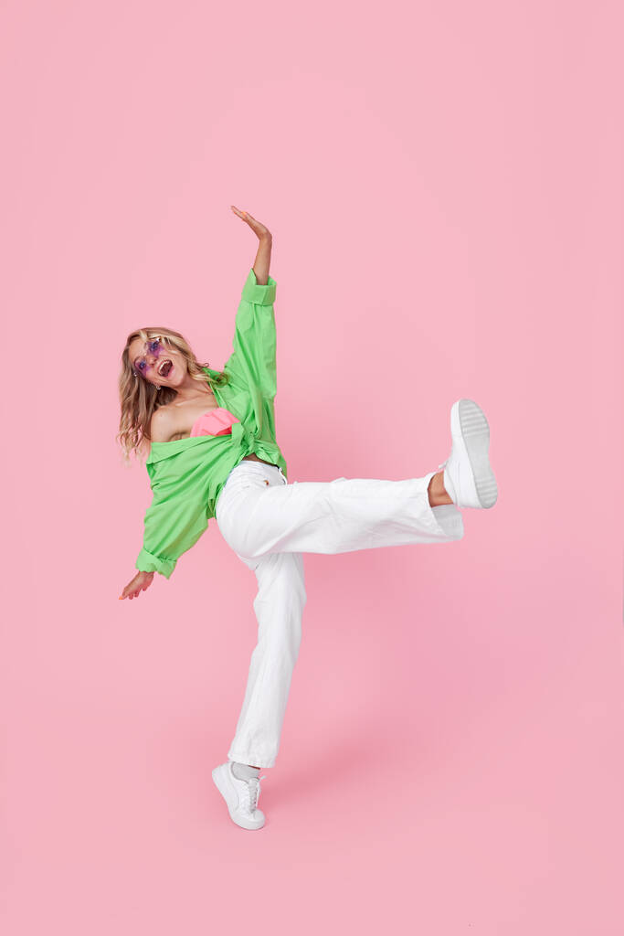 Full length view of pretty cheerful girl dancing in good mood in green bright shirt isolated on pink background - Photo, image