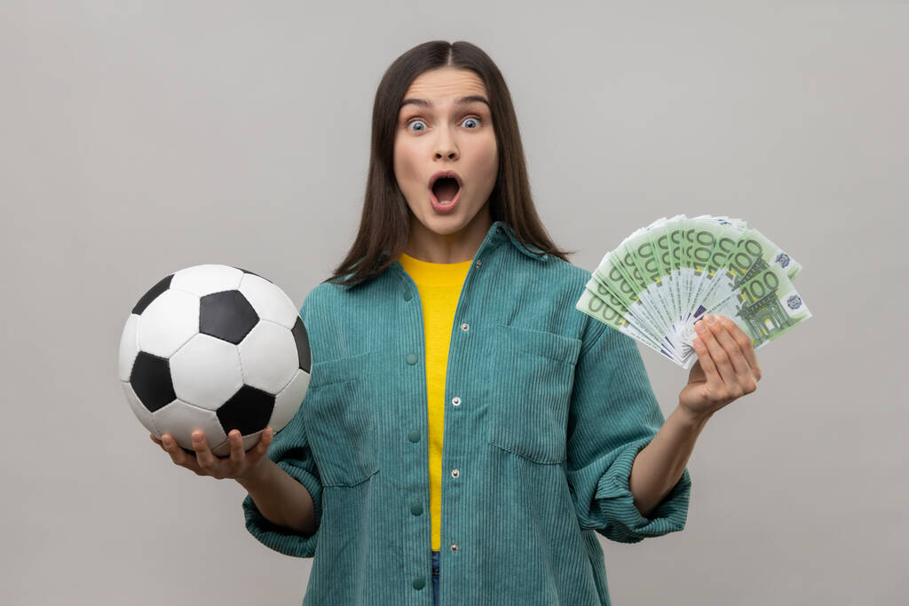 Shocked amazed woman showing soccer ball and fun of hundred euro bills, winning lot of money betting for sport, wearing casual style jacket. Indoor studio shot isolated on gray background. - Fotoğraf, Görsel