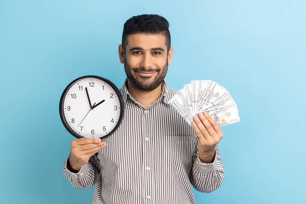 Positive handsome bearded businessman holding big fan of dollar banknotes and wall clock, time is money, expressing happiness, wearing striped shirt. Indoor studio shot isolated on blue background. - Φωτογραφία, εικόνα