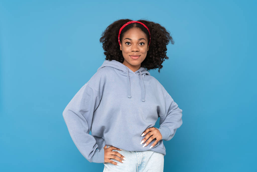 jonge zwarte vrouw dragen hoofdband glimlachen en kijken naar camera geïsoleerd over blauwe achtergrond - Foto, afbeelding