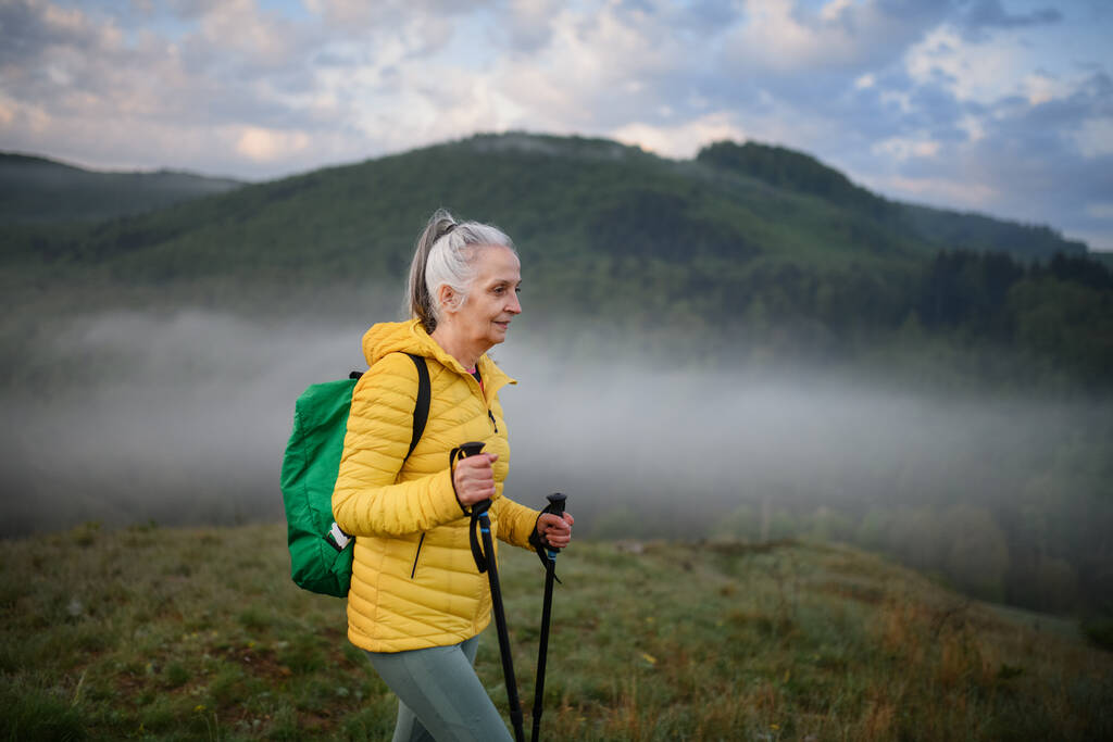 Une femme âgée randonnée dans la nature tôt le matin avec brouillard et montagnes en arrière-plan. - Photo, image