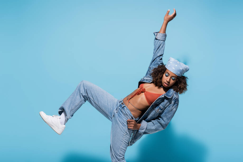 african american woman in denim clothes posing on one leg with arm up on blue background - Photo, Image
