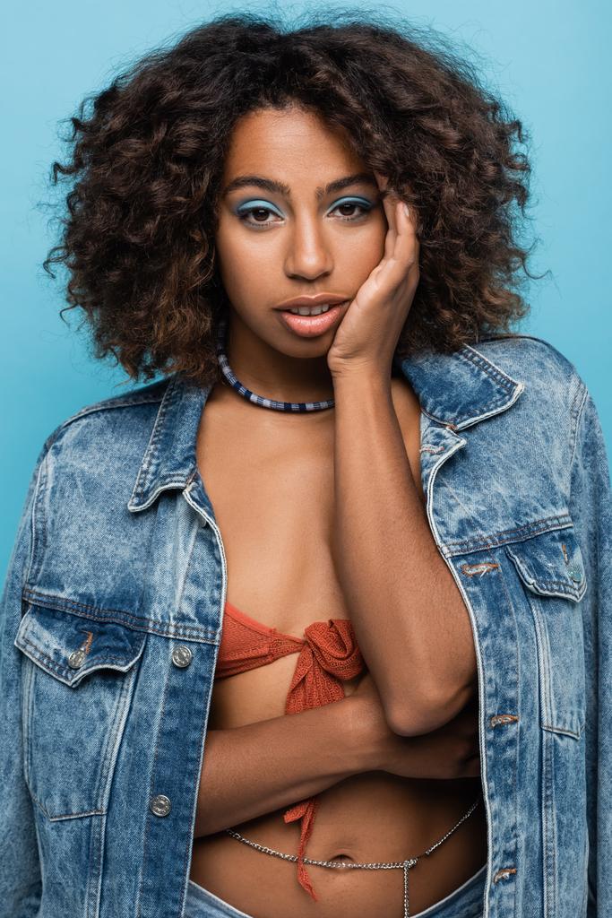 sensual african american woman in denim jacket touching face and looking at camera isolated on blue - Фото, изображение