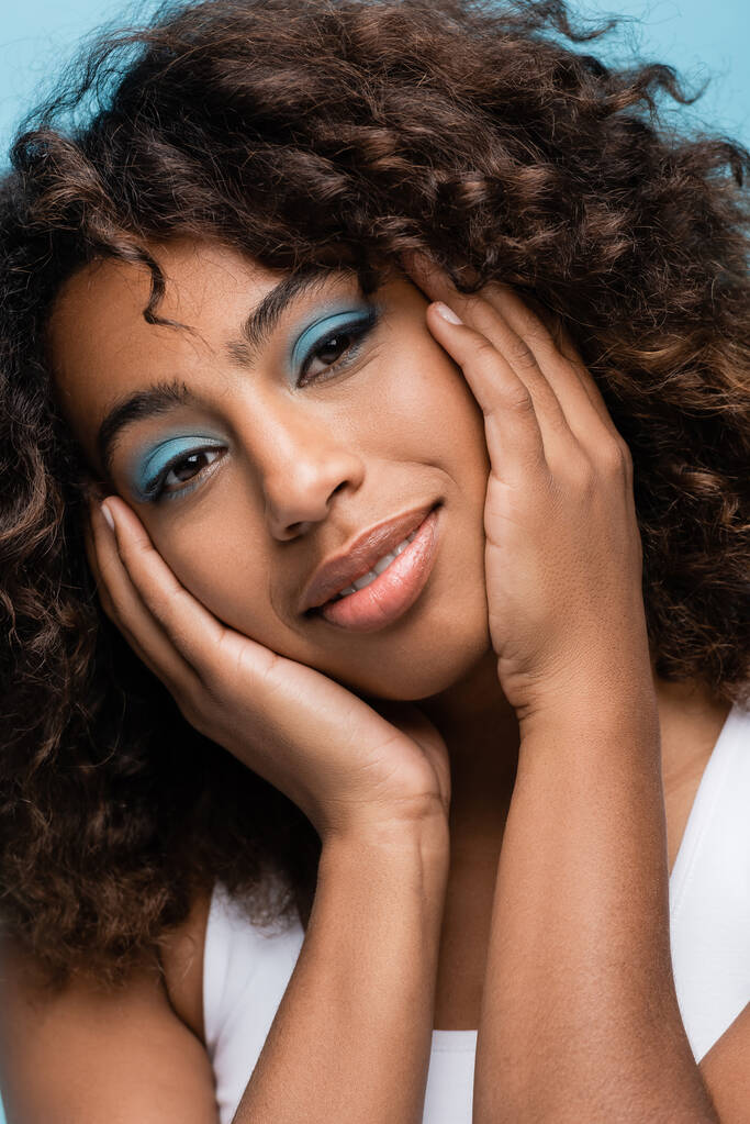 portrait of african american woman with makeup touching face and smiling at camera isolated on blue - Fotografie, Obrázek