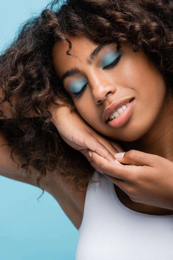 happy african american woman with closed eyes and makeup holding hands near perfect face isolated on blue - 写真・画像