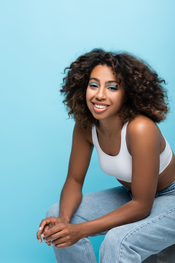 brunette african american woman in white crop top smiling at camera while sitting isolated on blue - Фото, изображение