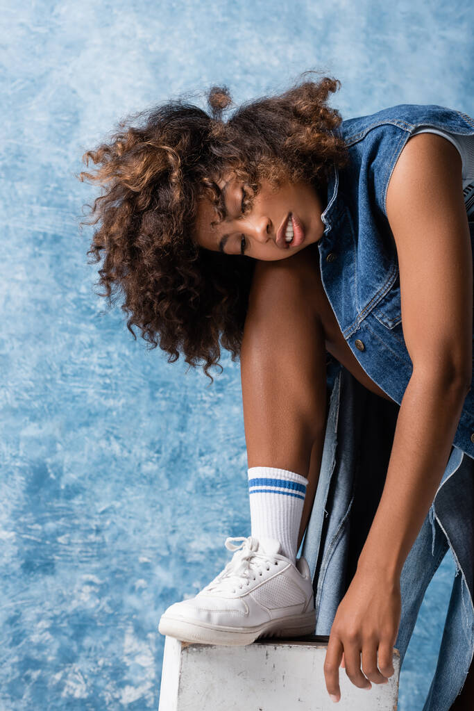 curly african american woman in denim clothes and white sneaker posing on blue background - Foto, Imagen